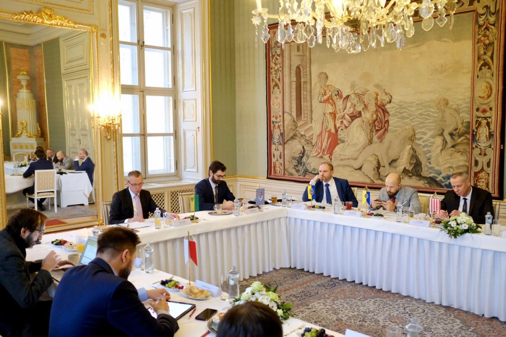 členovia združenia K8 sedia za stolom potiahnnutým bielym obrusom, vyzdobeným kvetmi, pred každým členom je vlajka mesta, ktoré zastupujú, stretnutie je v barokovej sieni, zrkadlo po ľavej strane a gobelín na pravej