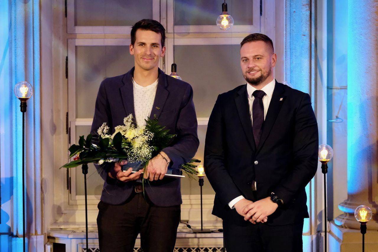 dvaja slušne oblečení muži stoja, muž  vľavo má v rukách kyticu a diplom, muž v pravo nemá nič