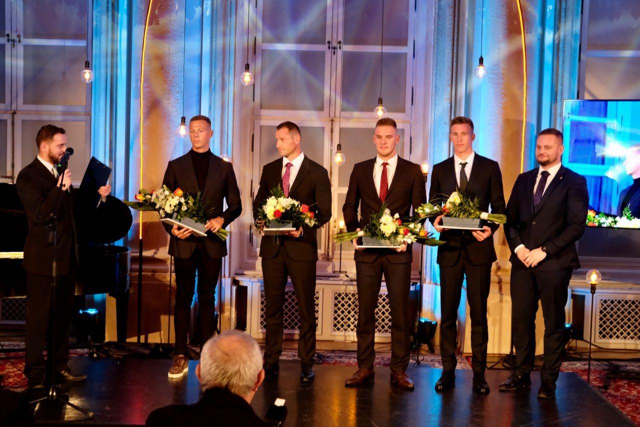 štyria slušne oblečení muži stoja s kyticami a diplomami, vedľa nich v ľavo stojí moderátor s mikrofónom na pravo stojí ďalší pekne oblečený muž